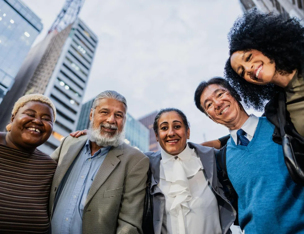 Grupo de pessoas de idades e perfis diferentes, reunidas e sorrindo numa rua da cidade